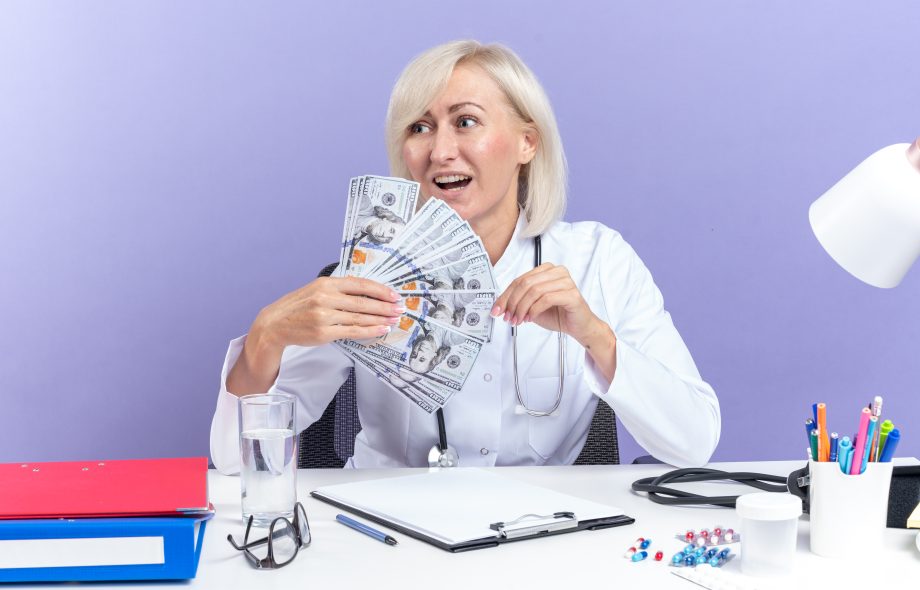 joyful-adult-slavic-female-doctor-medical-robe-with-stethoscope-sitting-desk-with-office-tools-holding-money-looking-side-isolated-purple-background